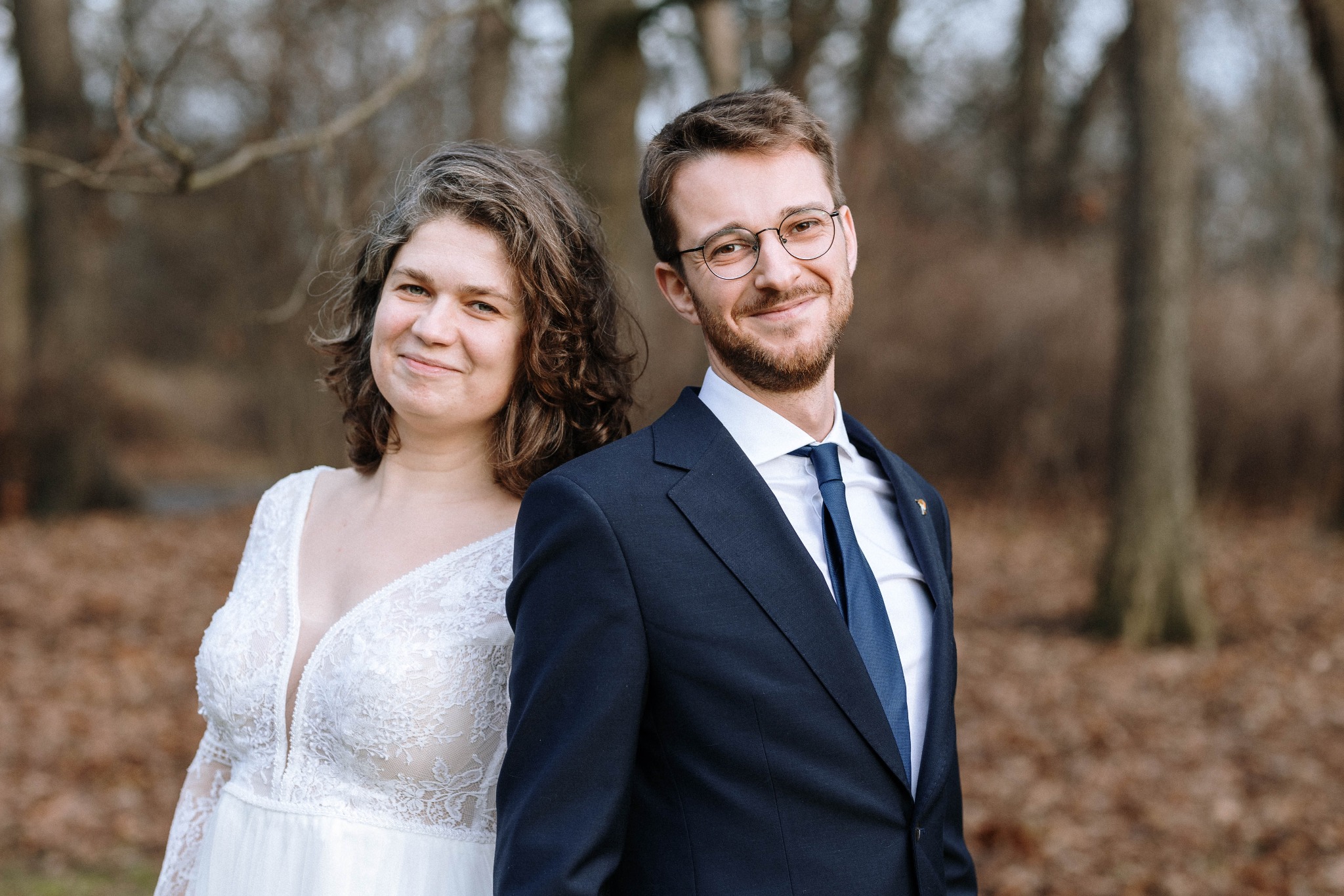 Para młoda: Panna młoda w koronkowej sukni z głębokim dekoltem, Pan młody w granatowym garniturze z niebieskim krawatem i okularach, stojący blisko siebie na tle jesiennego parku.