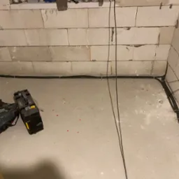 Surowy stan: czarne kable elektryczne ułożone wzdłuż ścian z betonu komórkowego, narzędzia i torba monterska na podłodze, widoczne puszki instalacyjne z przewodami.