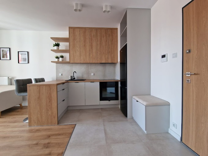 Nowoczesna kuchnia z drewnianymi szafkami górnymi i białymi dolnymi, czarny zlew i bateria, dwie półki z roślinami, oraz ława z tapicerowanym siedziskiem przy drzwiach.