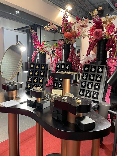 Ekspozycja biżuterii na czarnym stole z miedzianymi nogami, dekorowana kwiatami w ciemnych wazonach, z lustrem w tle.