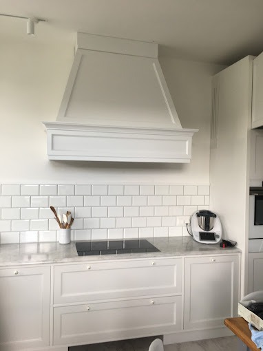 Biała kuchnia z okapem w stylu klasycznym, białe płytki na ścianie, marmurowy blat, białe szafki z frezowanymi frontami i okrągłymi gałkami, płyta indukcyjna.