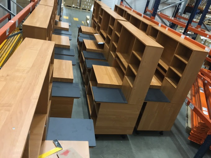 Magazyn z elementami drewnianych mebli modułowych z półkami, blaty w kolorze grafitowym, widoczne regały magazynowe w tle.