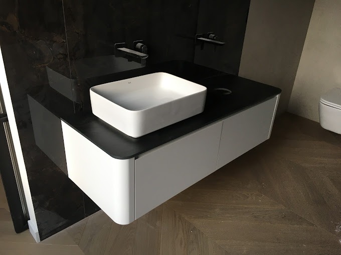 Nowoczesna łazienka z białą, prostokątną umywalką nablatową na czarnym blacie szafki z białymi, zaokrąglonymi frontami, ciemna ściana z marmuru w tle i parkiet w jodełkę na podłodze.