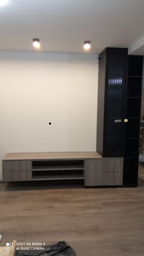 Nowoczesna zabudowa ściany w salonie: szara szafka RTV z półkami i czarna wysoka szafa z otwartymi półkami, podświetlenie sufitowe, panele podłogowe.