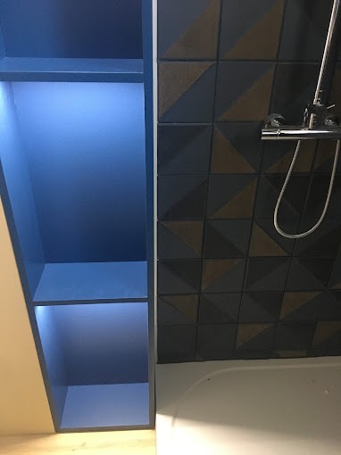 Niebieska, podświetlana szafka z półkami obok wanny z baterią i ciemnymi, geometrycznymi płytkami na ścianie.