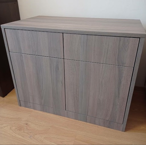 Niska, dwudrzwiowa szafka z szarego laminatu imitującego drewno, z prostymi frontami i subtelnymi uchwytami, na jasnej drewnianej podłodze.