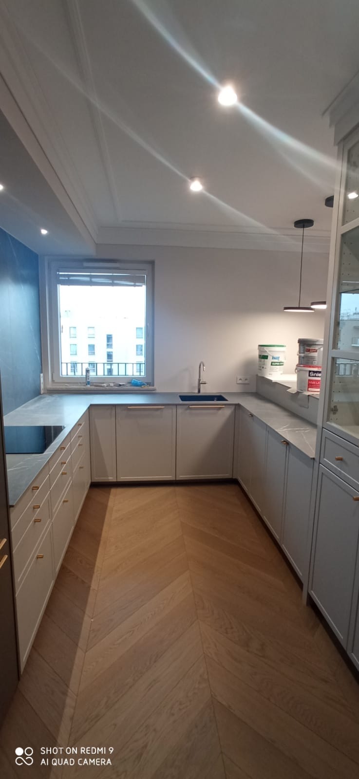 Jasna kuchnia w kształcie litery U z drewnianą podłogą w jodełkę, szarym blatem i widokiem na miasto z okna, uzupełniona farbami w tle.