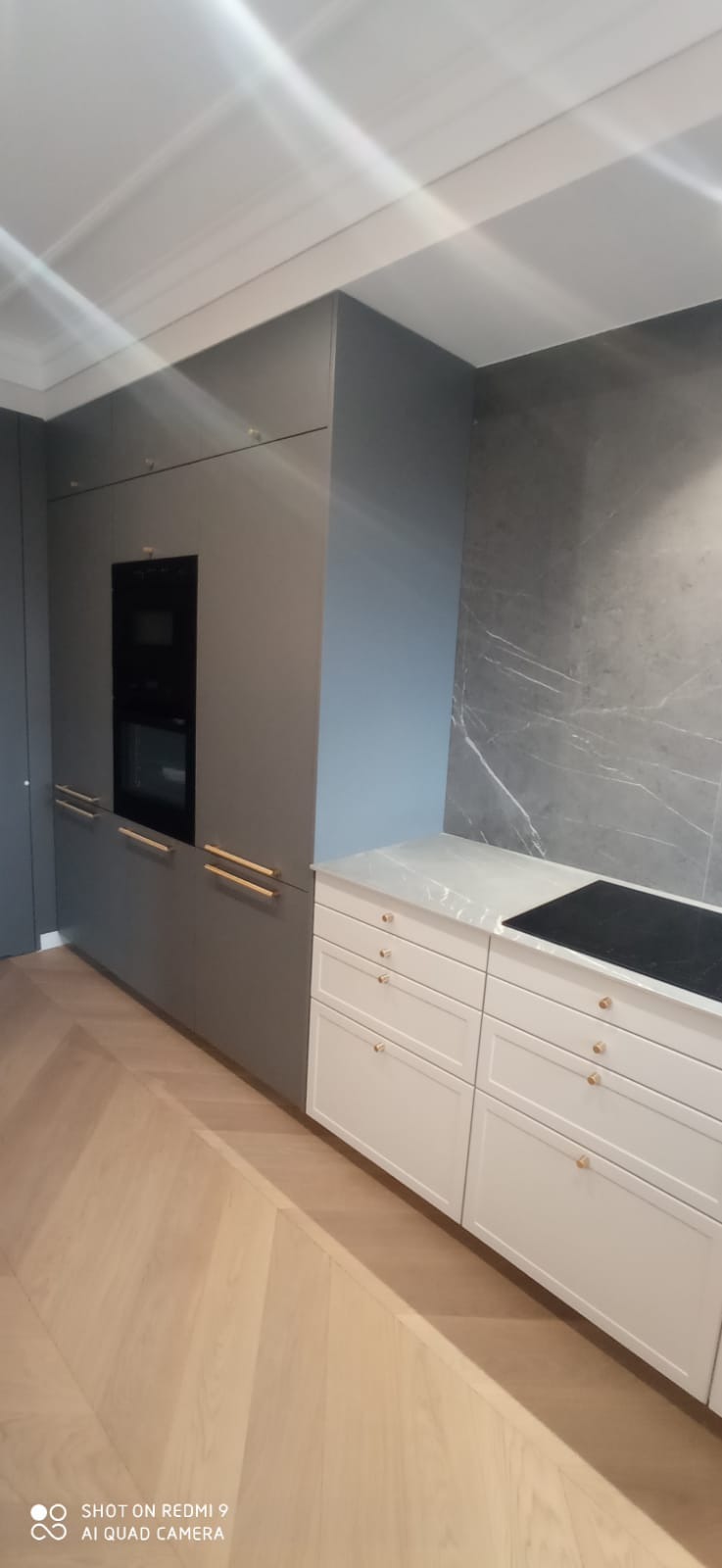 Nowoczesna zabudowa kuchenna z szarymi szafkami z wbudowanym piekarnikiem i białymi szafkami dolnymi z marmurowym blatem oraz płytą indukcyjną, na tle ściany z szarego marmuru.