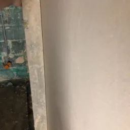 Gładka, jasna ściana po tynkowaniu maszynowym, widoczny fragment surowej ściany z instalacją elektryczną i fragment dłoni przy krawędzi tynku.