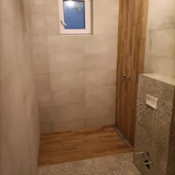 Wykończona łazienka z beżowymi płytkami ściennymi, drewnopodobnymi płytkami w strefie prysznica i na podłodze w kabinie oraz dekoracyjnymi płytkami podtynkowego WC.