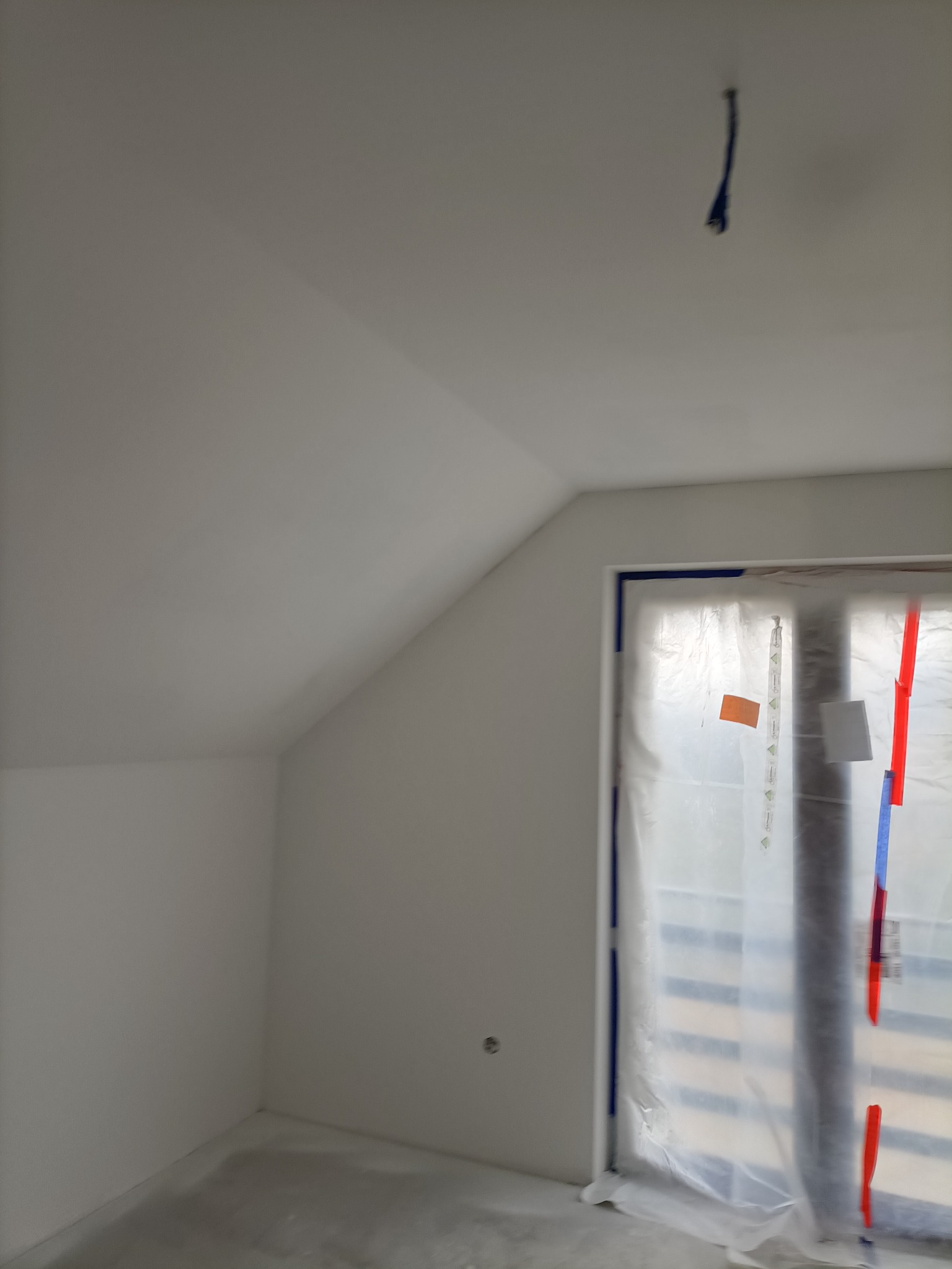 Poddasze po malowaniu na biało, okno zabezpieczone folią i taśmą malarską, widoczny kabel zwisający z sufitu.