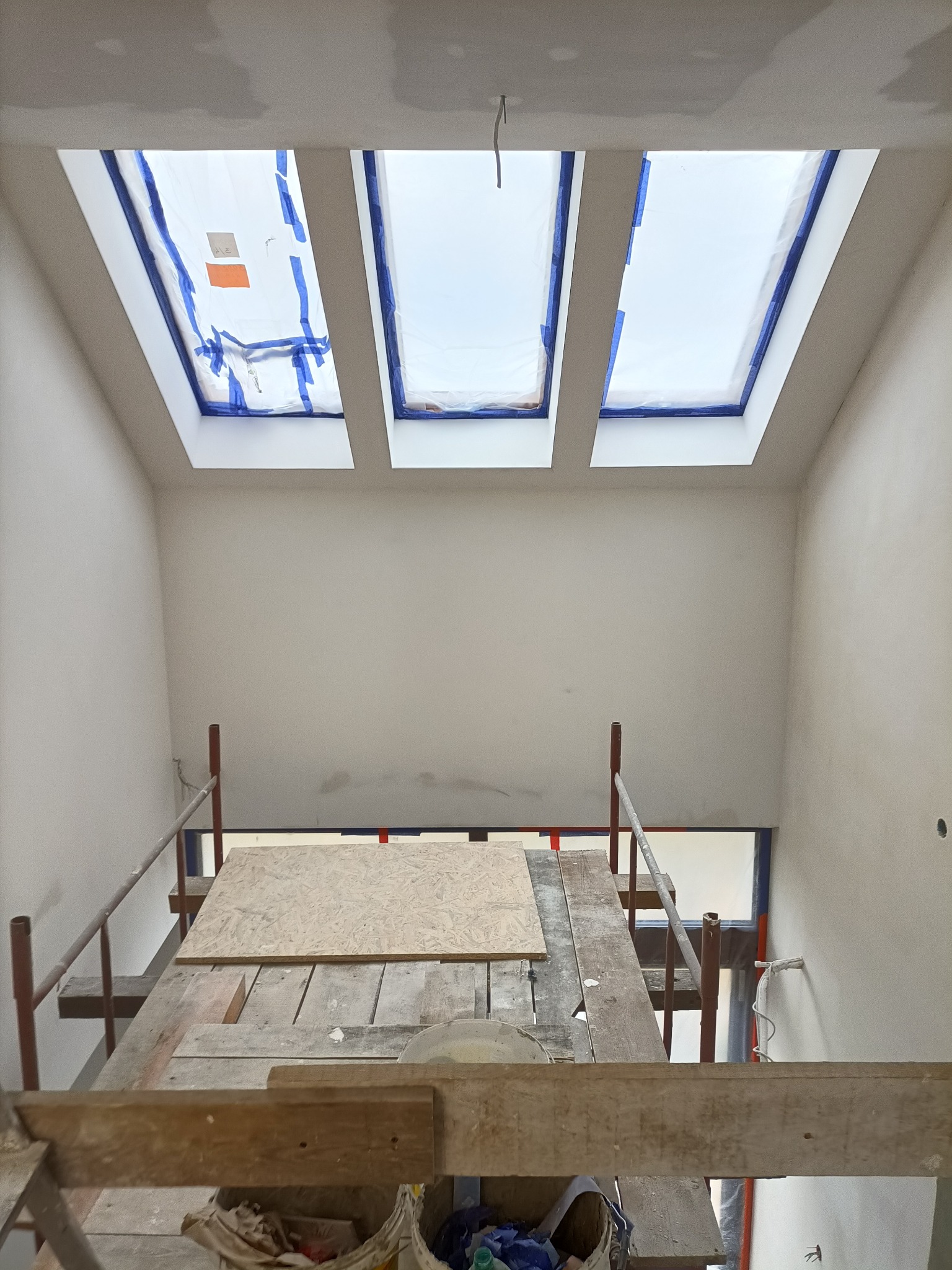 Wnętrze w trakcie remontu, widok z góry na rusztowanie i trzy okna dachowe zabezpieczone folią i taśmą malarską, ściany przygotowane do malowania.