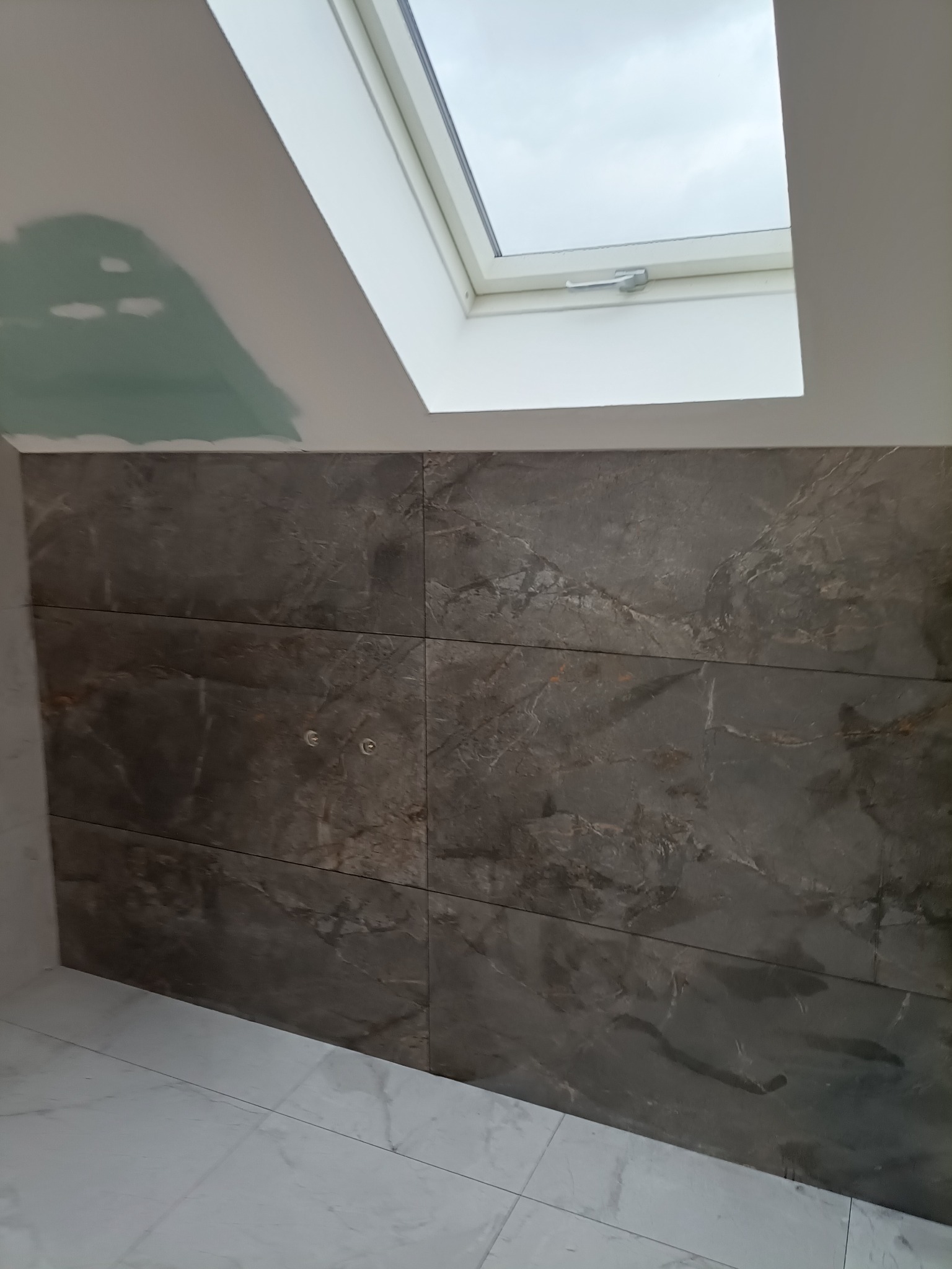 Ściana łazienki w trakcie wykańczania, z położonymi ciemnymi płytkami imitującymi marmur, widoczne otwory na armaturę, podłoga wyłożona jasnymi płytkami, okno dachowe i fragment sufitu z widocznymi...