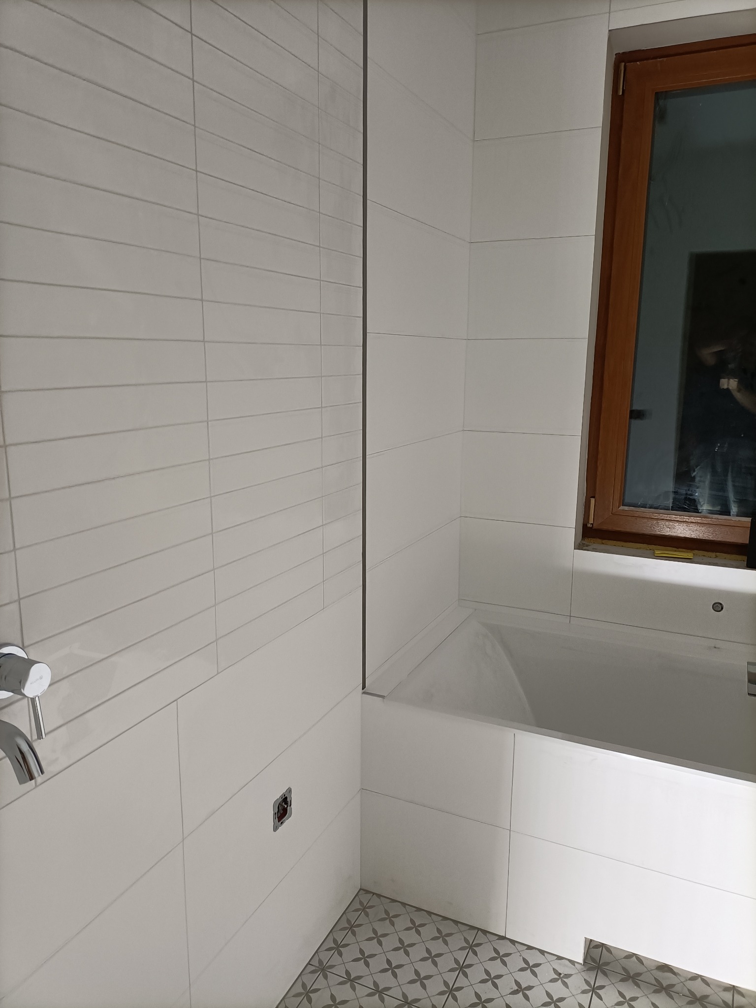 Wykończona łazienka z wanną obłożoną białymi, poziomymi płytkami, drewnianym oknem i wzorzystą podłogą; widoczny fragment baterii i otwór na gniazdko elektryczne.