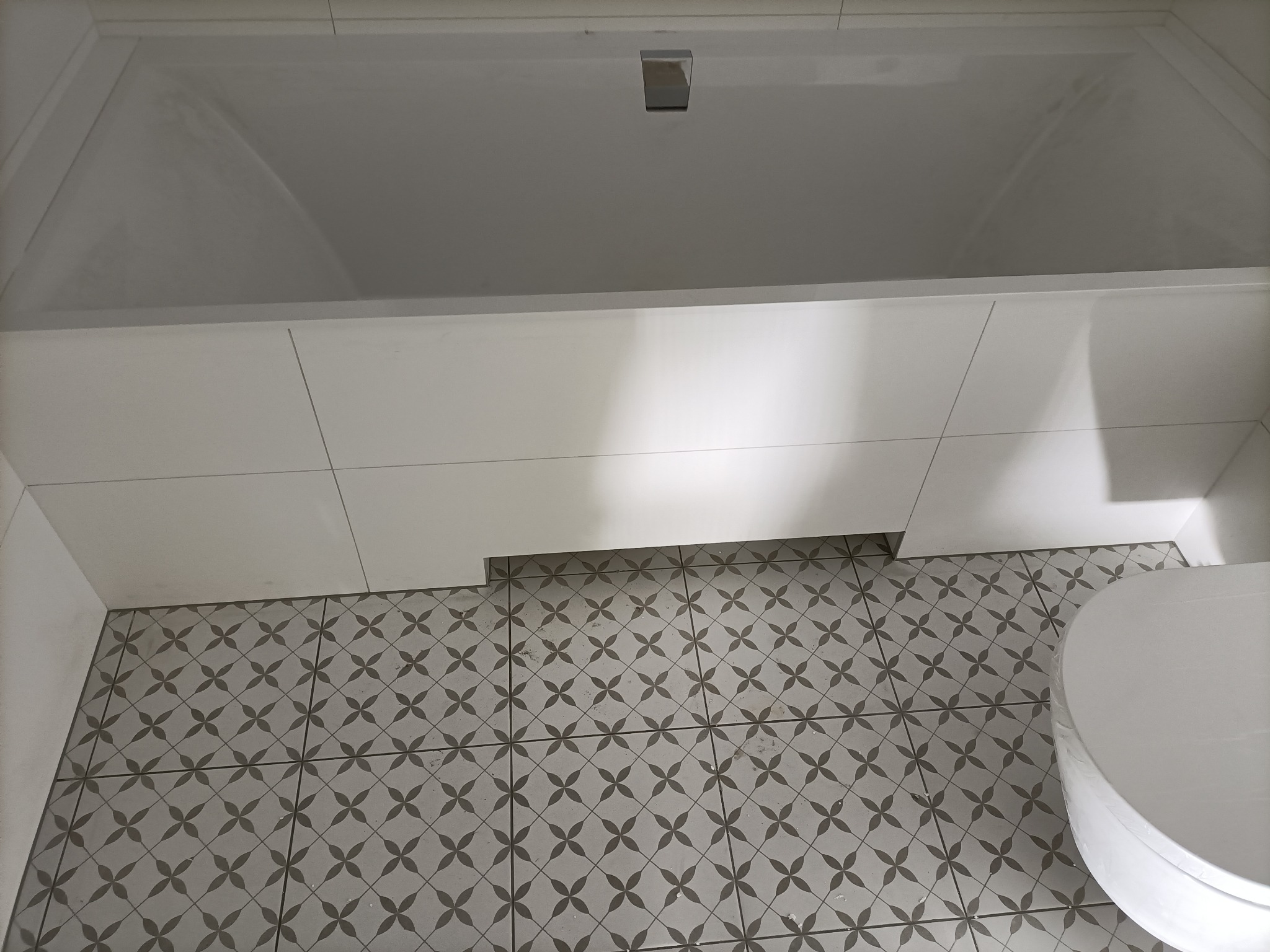 Nowoczesna, jasna łazienka z białą wanną obudowaną płytkami i szarą podłogą w geometryczny wzór kwiatowy.