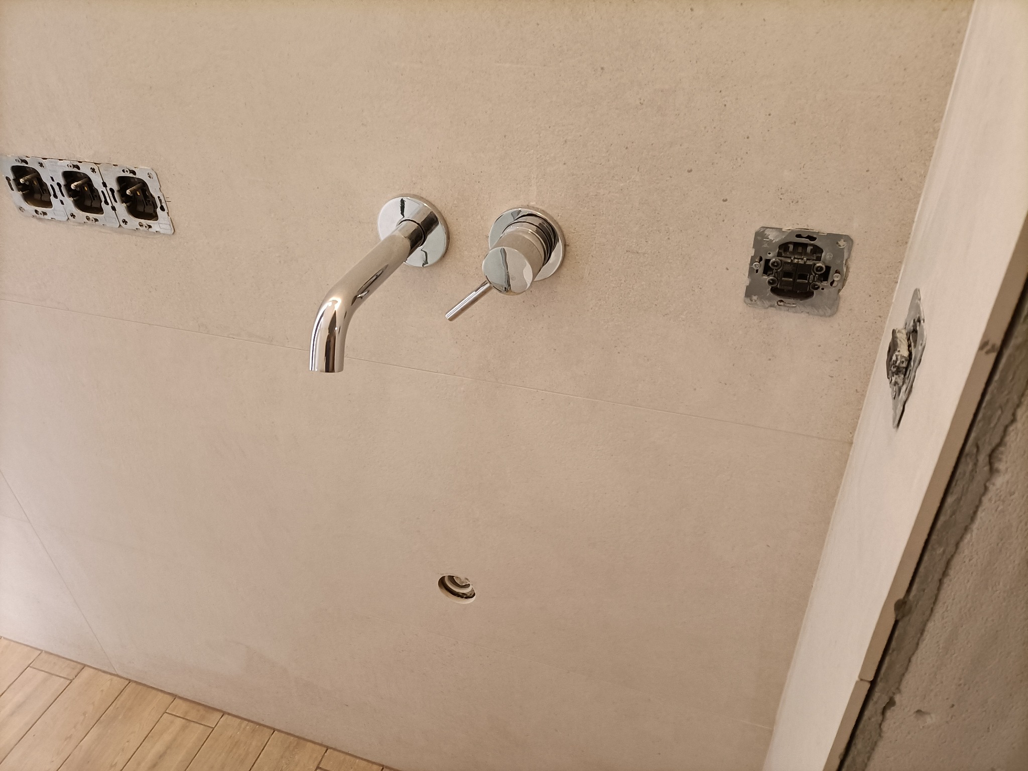 Ściana łazienki wyłożona jasnymi płytkami z zamontowaną chromowaną baterią umywalkową i przygotowanymi otworami na gniazdka elektryczne, widoczne drewnopodobne płytki podłogowe.