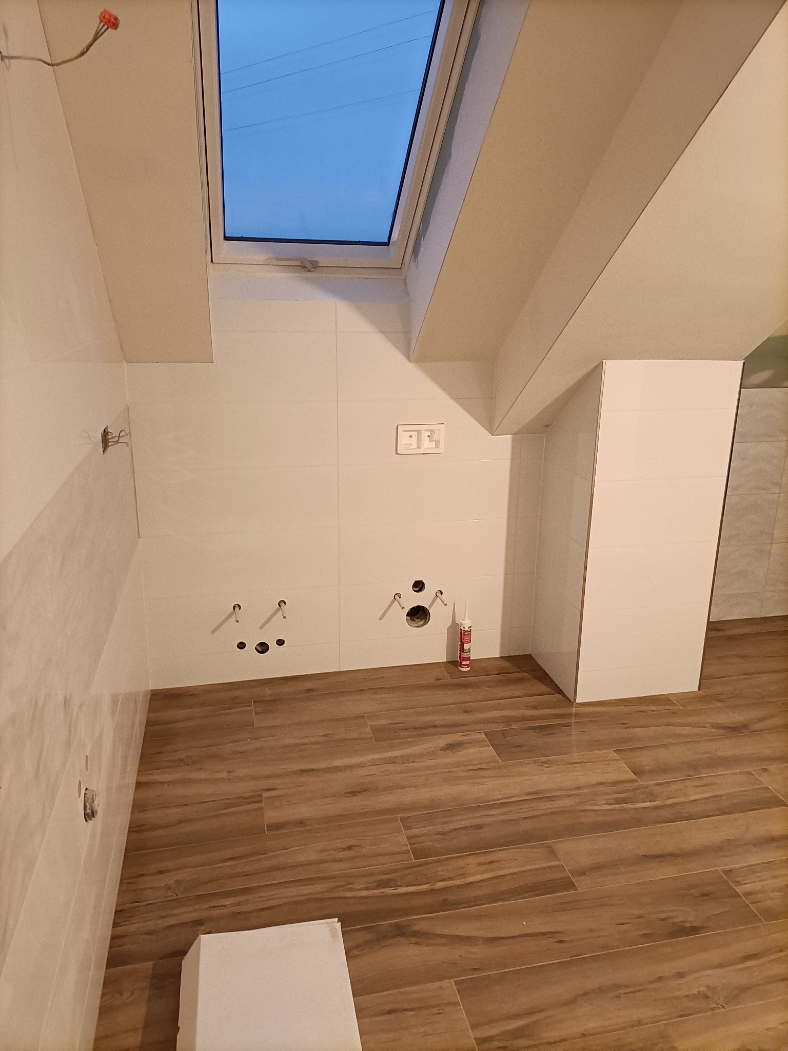 Wnętrze łazienki w trakcie remontu z drewnopodobną podłogą, białe płytki na ścianach, okno dachowe i widoczne przyłącza wodno-kanalizacyjne oraz tuba silikonu sanitarnego.