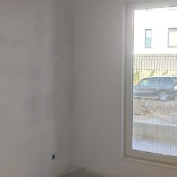 KADRA SPÓŁKA Z OGRANICZONĄ ODPOWIEDZIALNOŚCIĄ - Pokój w trakcie prac wykończeniowych: ściany pokryte szpachlą, widoczne przewody elektryczne wystające z sufitu, okno z widokiem na zewnątrz.