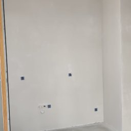 KADRA SPÓŁKA Z OGRANICZONĄ ODPOWIEDZIALNOŚCIĄ - Wnętrze w trakcie prac wykończeniowych: ściany pokryte świeżą warstwą, widoczne puszki z masą szpachlową, przewody elektryczne wystające z sufitu i drabina oparta o ścianę.