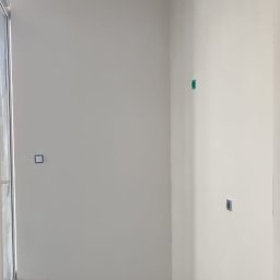 KADRA SPÓŁKA Z OGRANICZONĄ ODPOWIEDZIALNOŚCIĄ - Przygotowane do malowania, wyszpachlowane ściany w pomieszczeniu z widocznymi przewodami elektrycznymi wystającymi z sufitu i mikserem do gładzi na podłodze.