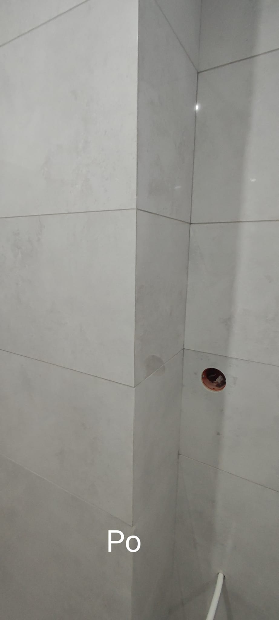 Wykończony narożnik łazienki z jasnymi, prostokątnymi płytkami ceramicznymi, widoczny otwór na instalację elektryczną i fragment rury przy podłodze.