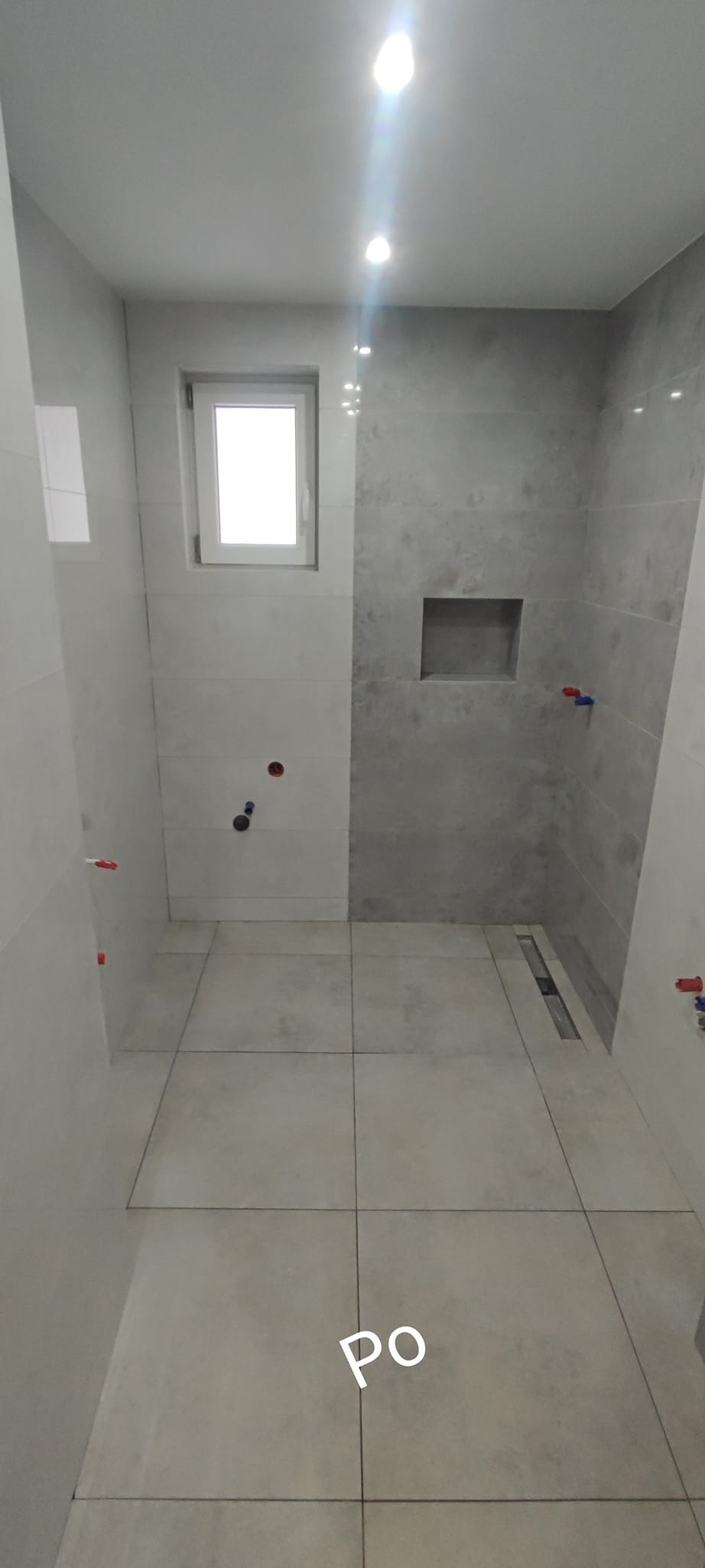 Wykończona łazienka z szarymi i białymi płytkami, widoczny odpływ liniowy, wnęka w ścianie i okno.