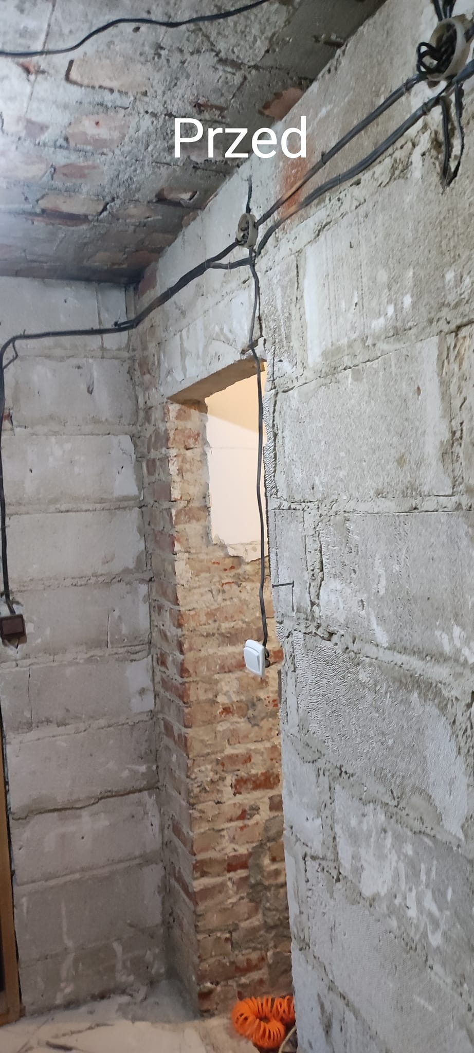 Surowy stan wnętrza pomieszczenia w trakcie remontu: ściany z betonu komórkowego i cegły, widoczne kable elektryczne, brak wykończenia, napis 'Przed' sugerujący etap przed pracami.