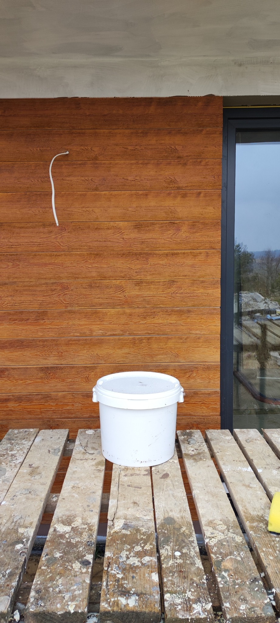 Białe wiadro na drewnianym podeście przed ścianą z paneli imitujących drewno, obok okno z widokiem na zewnątrz.