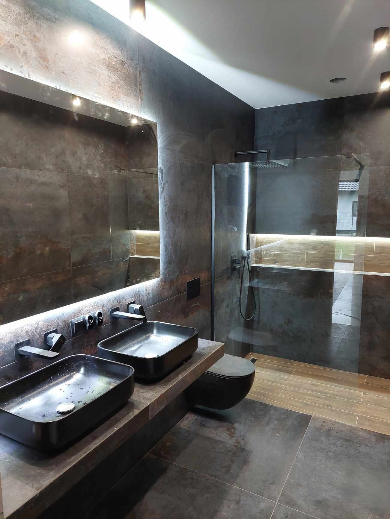 Nowoczesna łazienka z dwoma czarnymi umywalkami nablatowymi, lustrem z podświetleniem LED, kabiną prysznicową typu walk-in i ciemnymi płytkami imitującymi beton.