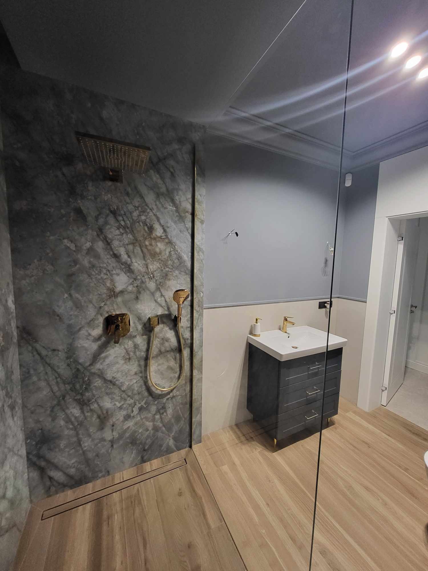 Nowoczesna łazienka z marmurową ścianą prysznicową, złotą armaturą i drewnopodobną podłogą, widok na umywalkę z szufladami i szklaną ścianę kabiny.