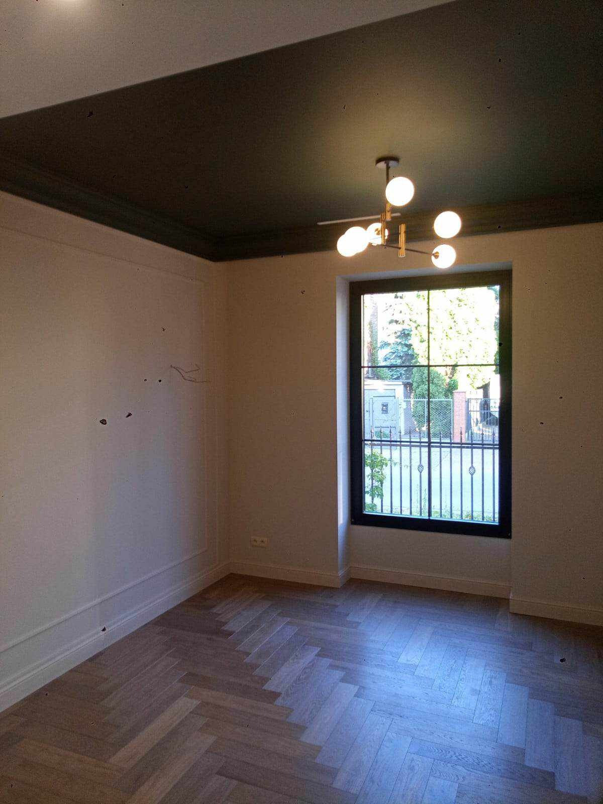 Pokój po częściowym remoncie: parkiet ułożony w jodełkę, pomalowany na ciemno sufit z dekoracyjną listwą, okno z czarną ramą i widokiem na zieleń, ściany przygotowane do malowania z widocznymi...