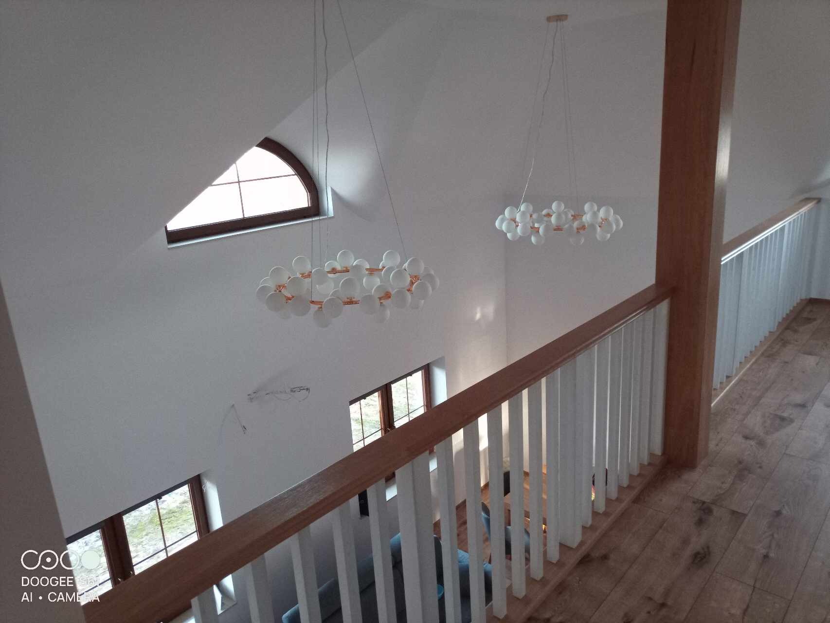 Wnętrze domu po remoncie, widok z góry na białe ściany, drewnianą podłogę, białą balustradę z drewnianym poręczą oraz nowoczesne lampy wiszące w kształcie kuli.