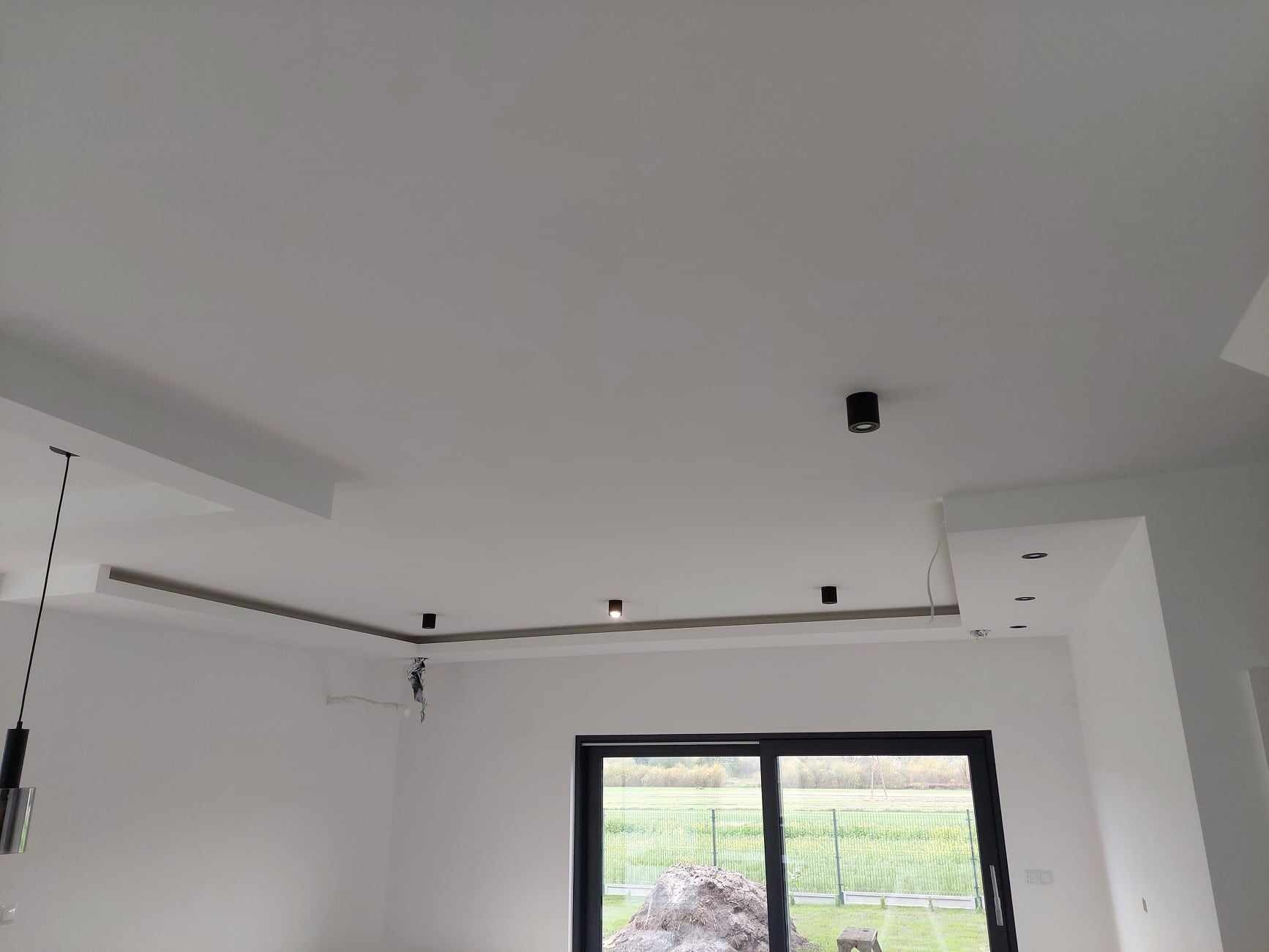 Białe wnętrze po remoncie z zamontowanymi czarnymi lampami sufitowymi, widoczne wystające kable i niedoskonałości na ścianie, okno z widokiem na zewnątrz.