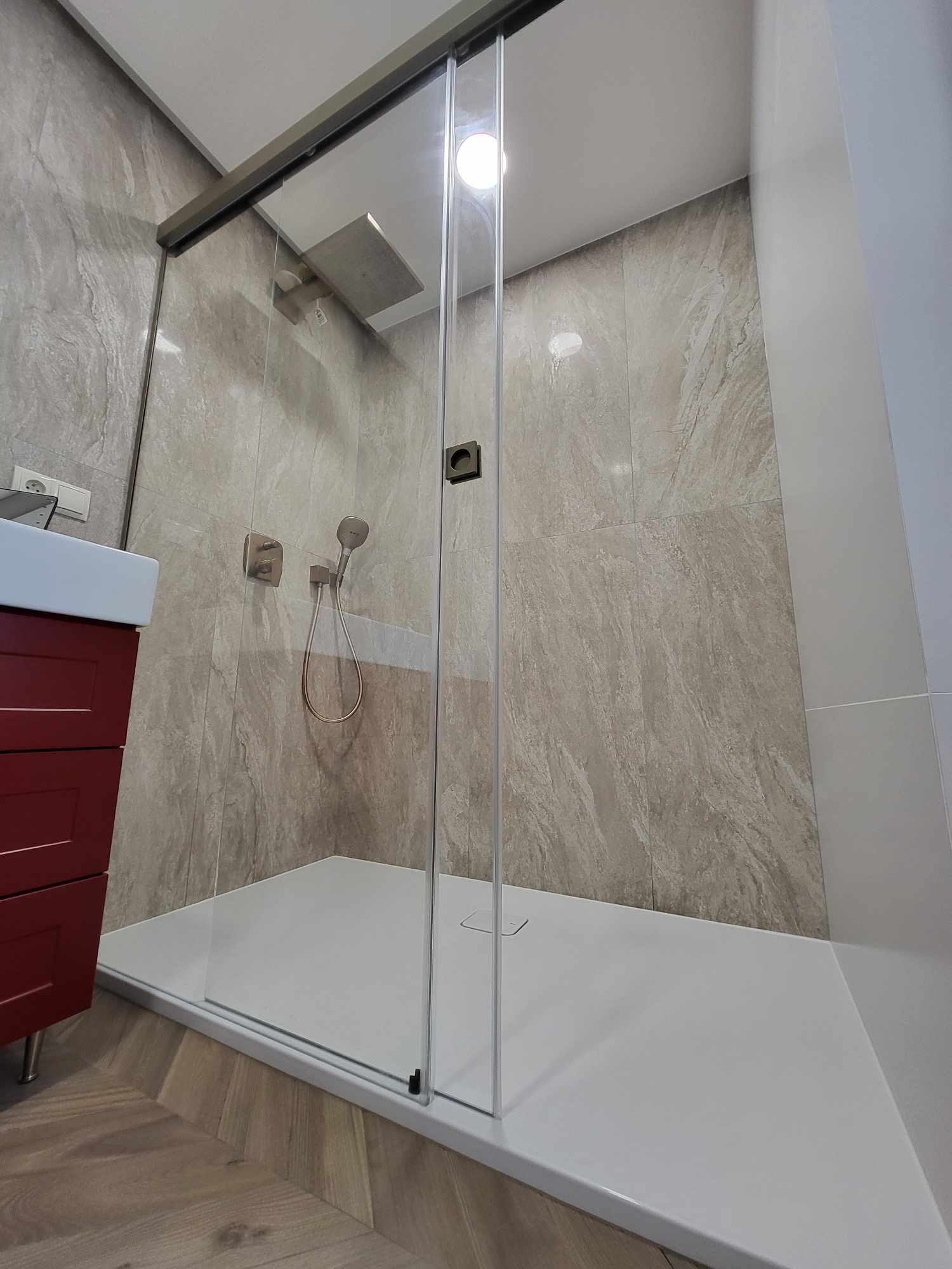Nowoczesna kabina prysznicowa z przesuwnymi drzwiami, wykończona płytkami imitującymi kamień, z widocznym panelem prysznicowym i słuchawką, oraz czerwoną szafką pod umywalką.