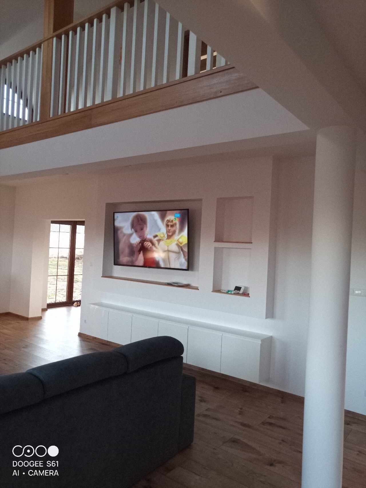 Jasne wnętrze domu z antresolą, drewnianą podłogą i zabudową meblową pod telewizorem w ścianie.