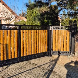 MAJMETAL - Nowoczesna brama wjazdowa z drewnianymi panelami i czarną, metalową ramą z motywem greckim, na tle domu jednorodzinnego i drzew, z widocznym cieniem osoby fotografującej.