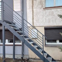 MAJMETAL - Zewnętrzne schody metalowe z balustradą, prowadzące do drzwi na piętrze, pomalowane na szaro, z widocznymi nitowanymi stopniami i prostą konstrukcją.