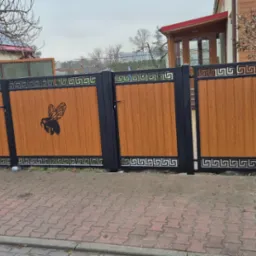 Nowoczesna brama wjazdowa z drewnopodobnym wypełnieniem i czarną ramą, ozdobiona motywem pszczoły i greckim wzorem, na tle domu jednorodzinnego.