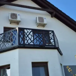 Czarna, ażurowa balustrada balkonowa o geometrycznym wzorze, zamontowana na białym budynku z dwoma klimatyzatorami i drabiną opartą o ścianę.
