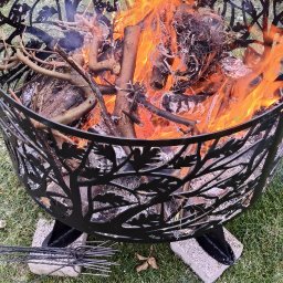 MAJMETAL - Metalowy kosz paleniskowy z wyciętym motywem drzewa, wypełniony płonącym drewnem, stoi na trawie. Widoczne narzędzia do ogniska.
