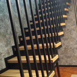 MAJMETAL - Nowoczesne schody z drewnianymi stopniami i minimalistyczną, metalową balustradą z pionowych prętów. Widok z dołu, podkreślający geometrię konstrukcji.