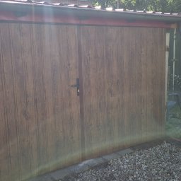 MAJMETAL - Dwuskrzydłowa brama garażowa z brązowymi panelami imitującymi drewno, z czarną klamką na tle zieleni i szarej kostki brukowej.