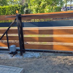 MAJMETAL - Nowoczesna brama przesuwna z drewnopodobnymi panelami i czarną ramą, widoczny napęd elektryczny. Brama zamontowana, otoczenie w trakcie prac wykończeniowych.