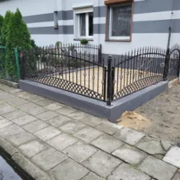 Ozdobne, niskie, czarne ogrodzenie z metalu, z motywem kratki i kwiatów, otaczające niewielki ogródek przed domem z szaro-białymi pasami, widok z boku, z fragmentem chodnika i żywopłotu.