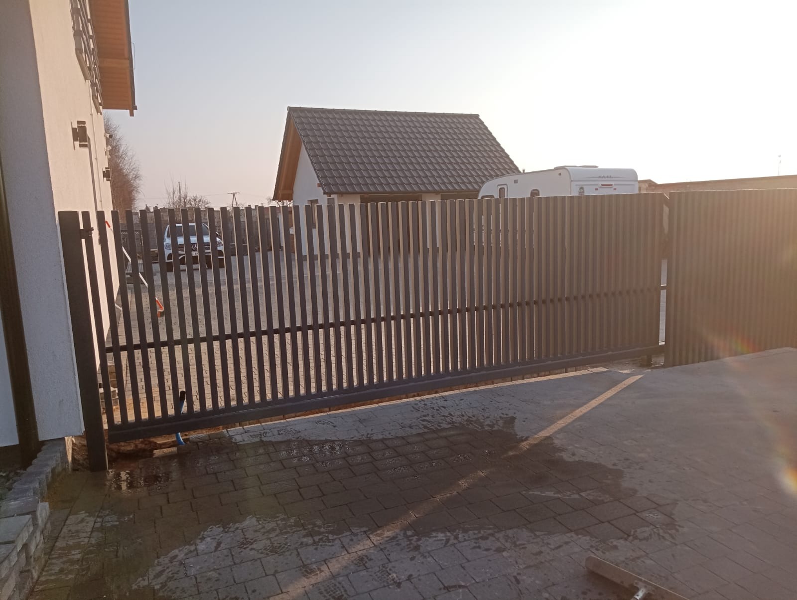 Nowoczesna, przesuwna brama wjazdowa z pionowych profili metalowych w kolorze antracytowym, widoczna na tle domu jednorodzinnego i przyczepy kempingowej w słoneczny dzień.