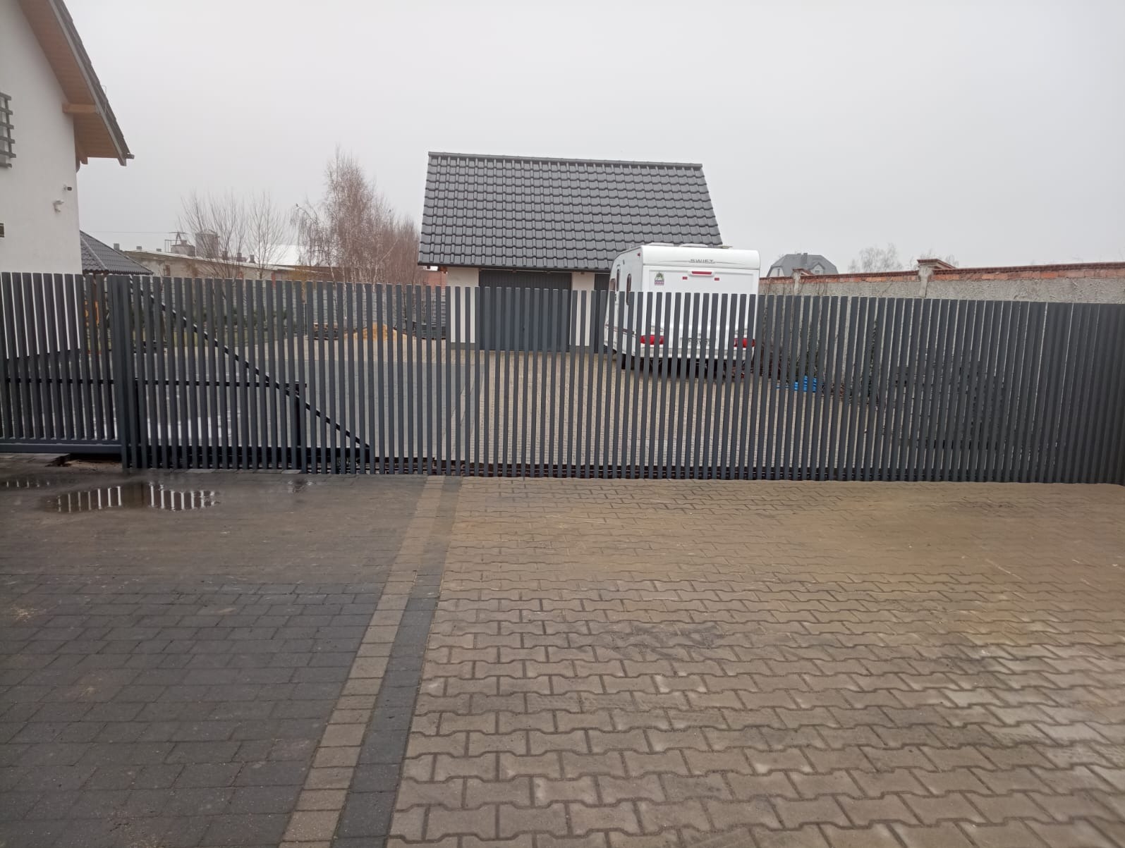 Nowoczesna, przesuwna brama wjazdowa z pionowych, szarych profili, z widocznym fragmentem posesji i przyczepą kempingową w tle.