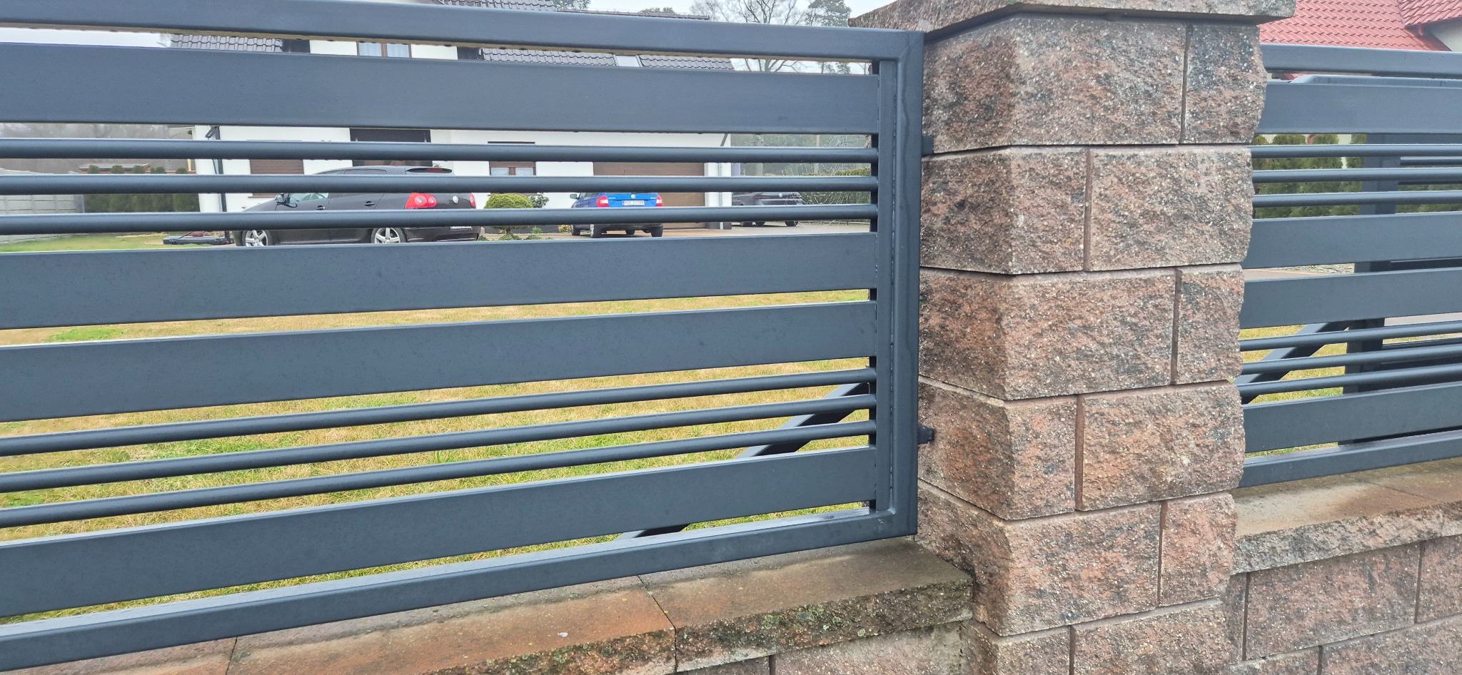 Nowoczesna brama wjazdowa z poziomych, szarych profili stalowych, osadzona na słupkach z brązowego kamienia łupanego, widok na podjazd z zaparkowanymi samochodami.