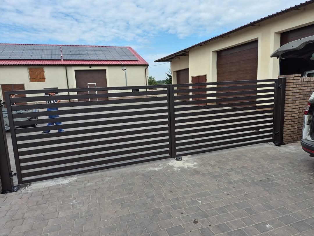 Nowoczesna, pozioma brama wjazdowa z metalowymi profilami w kolorze antracytowym, na tle budynku z panelami słonecznymi i garażami.