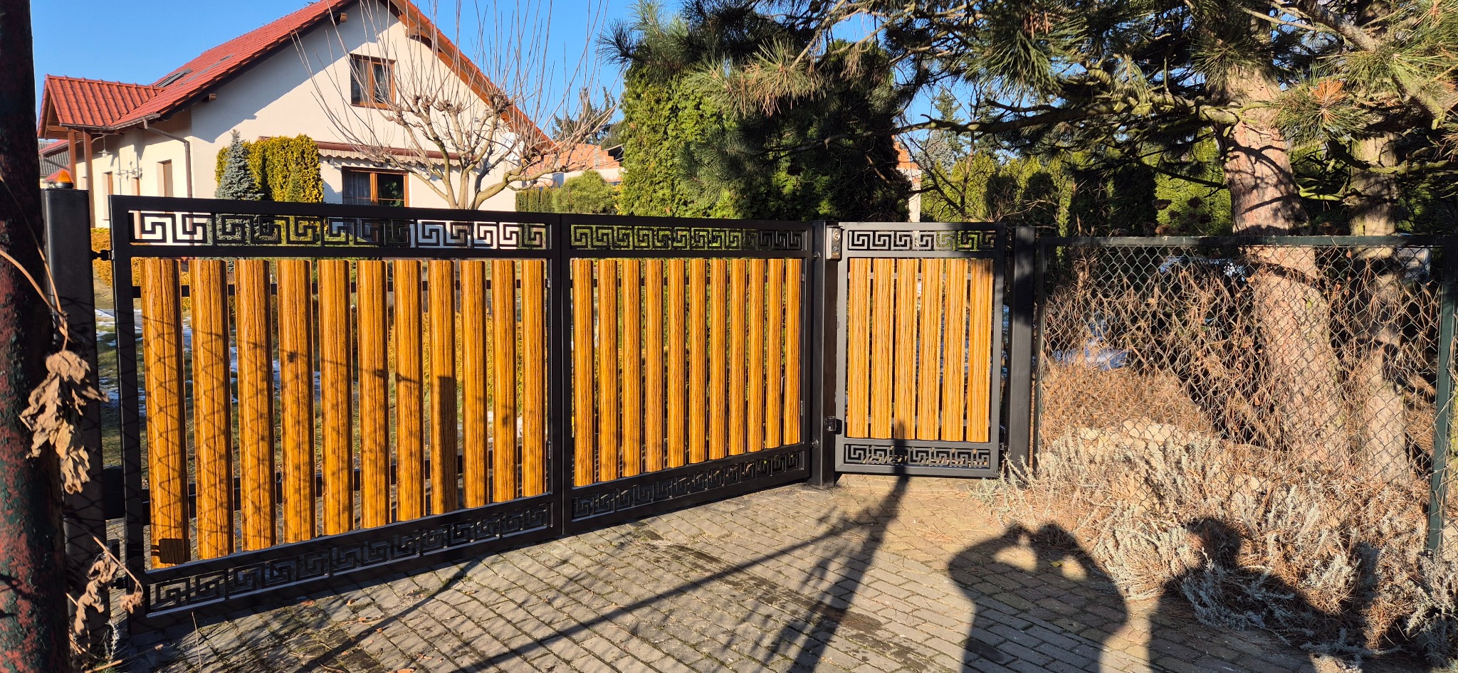 Nowoczesna brama wjazdowa z drewnianymi panelami i czarną, metalową ramą z motywem greckim, na tle domu jednorodzinnego i drzew, z widocznym cieniem osoby fotografującej.
