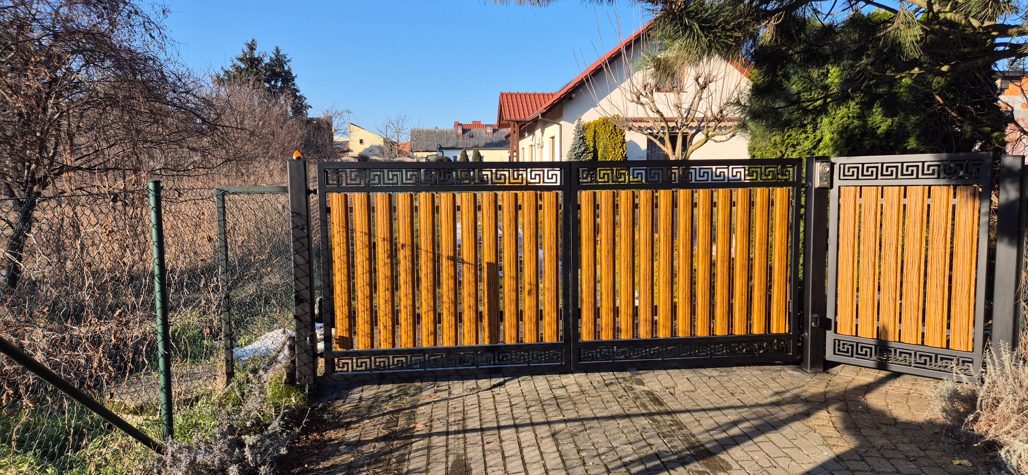 Dwuskrzydłowa brama wjazdowa z drewnianymi panelami i czarną metalową ramą ozdobioną greckim wzorem, na tle kostki brukowej i ogrodu.